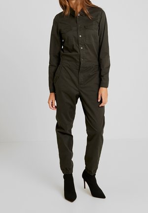 Combinaison en olive avec des manches longues, boutons à l'avant, deux poches poitrine, poignets élastiques aux chevilles, assortie à des bottines à talons hauts noires.