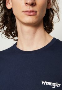 Temno modra bombažna majica z okroglim izrezom in kratkimi rokavi, z belim napisom "Wrangler" na levi strani prsnega dela.