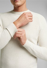 Homme ajustant le poignet d'un pull texturé à manches longues de couleur crème sur un fond gris uni.