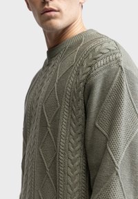 Olive green knitted sweater featuring a mix of textured patterns, including cable knit and diamond shapes, with a crew neck design.