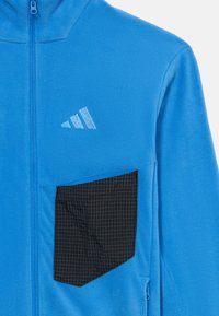 Μπλε φλις μπουφάν με ψηλό γιακά, που διαθέτει μαύρη τσέπη με σχέδιο πλέγματος και ανάγλυφο λογότυπο στο στήθος.