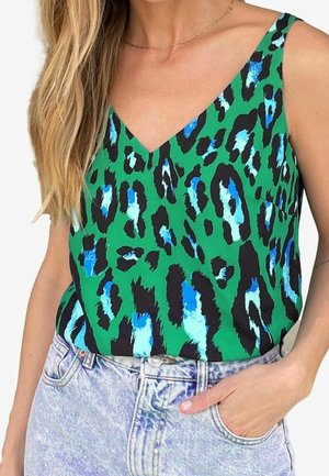 Kobieta w zielonym topie bez rękawów z niebiesko-czarnym nadrukiem w cętki lamparta i jasno-niebieskich jeansach z wysokim stanem, ręka w kieszeni.