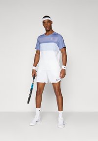 Tennisantrekk med en gradient blå og hvit t-skjorte med kort erme og hvite shorts, kombinert med hvite joggesko og et svart racket.