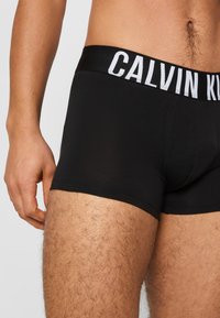 Black cotton boxer briefs with a wide white waistband featuring "CALVIN KLEIN" logo. Smooth texture, fitted design, and mid-thigh length.
