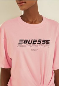 Camiseta de algodón rosa con un diseño gráfico negro que incluye el texto "GUESS" y "LOS ÁNGELES CALIFORNIA" debajo. Con mangas cortas y corte relajado.