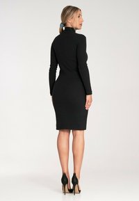 Czarna, dopasowana sukienka z długimi rękawami i wysokim kołnierzem. Gładki, elastyczny materiał sięga do połowy łydki. Noszona z czarnymi szpilkami.