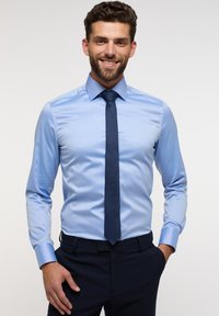 Chemise de couleur bleu clair avec une texture lisse, manches longues et col pointu. Assortie à une cravate à motifs foncés et un pantalon sombre.