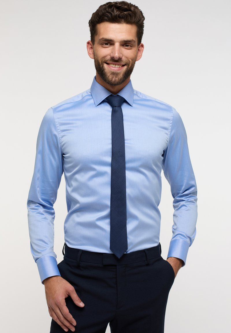 Chemise de couleur bleu clair avec une texture lisse, manches longues et col pointu. Assortie à une cravate à motifs foncés et un pantalon sombre.