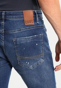Džíny v tmavomodrej farbe so slabým efektom vyblednutia, s hnedou koženou nášivkou označenou "CARS" a zadnou vreckom s bielymi škvrnami od farby.