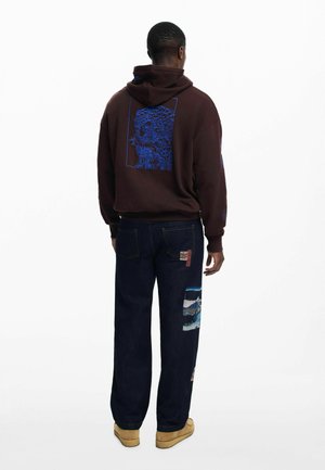 Sudadera marrón con un diseño gráfico azul en la parte posterior, combinada con jeans de mezclilla oscura que presentan detalles de remiendo y zapatos marrones claros.