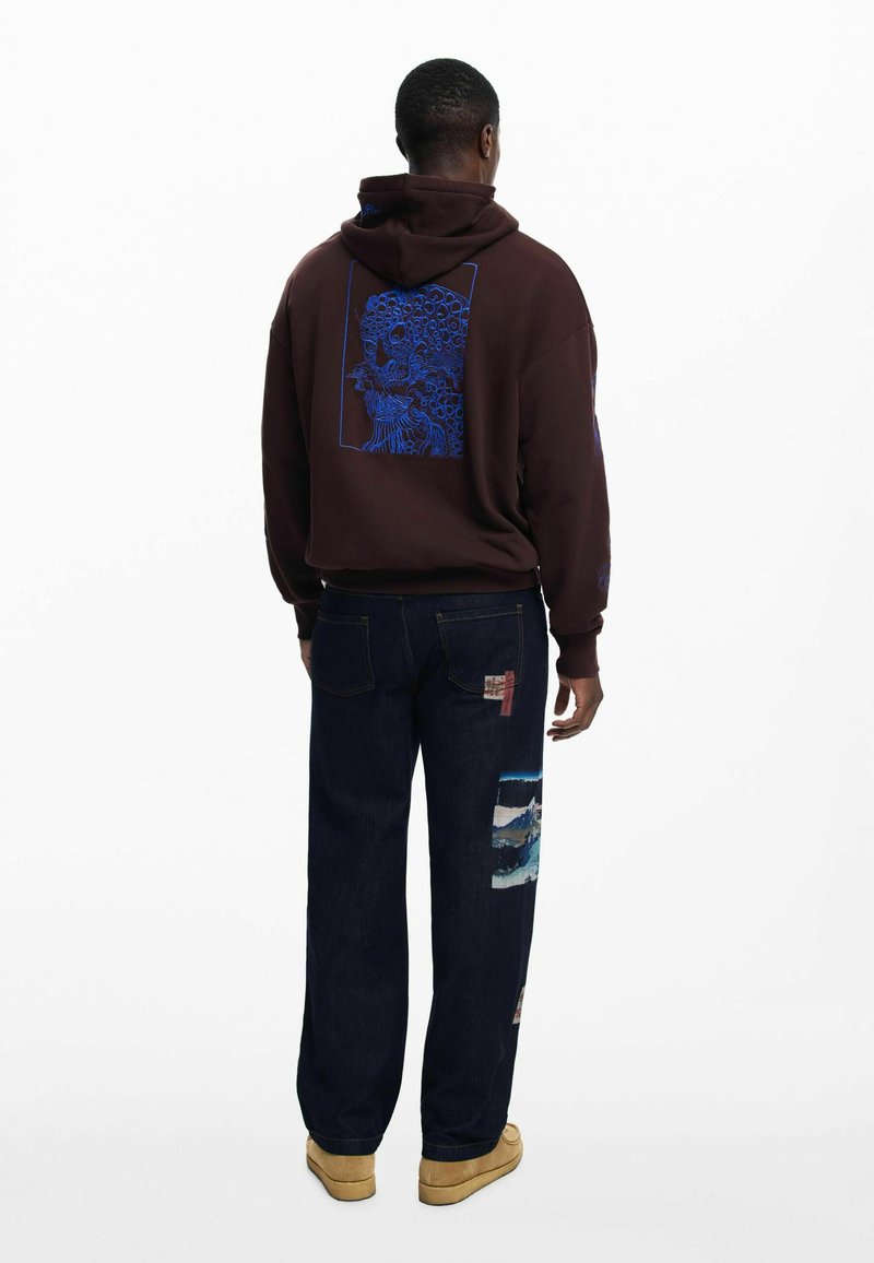 Brauner Kapuzenpullover mit blauem Grafikdesign auf dem Rücken, kombiniert mit dunkelblauen Jeans mit Patchwork-Details und hellbraunen Schuhen.