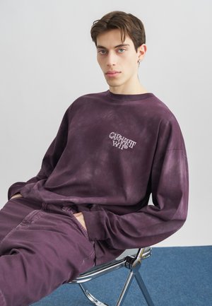 Młody mężczyzna o krótkich brązowych włosach, ubrany w fioletową bluzę z efektem batiku i dopasowane spodnie, siedzi na nowoczesnym metalowym krześle na tle białej ściany.