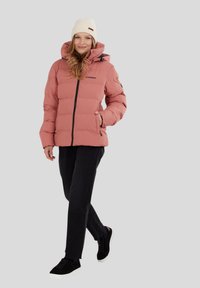Chaqueta de plumas rosa con cremallera frontal, cuello alto y puños elásticos, combinada con pantalones negros y zapatillas negras. Lleva un gorro de punto ligero en la cabeza.
