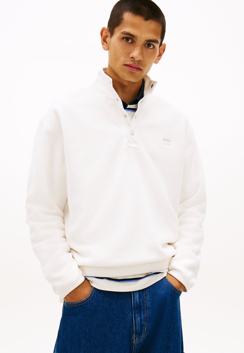 Άσπρο φλις πουλόβερ με μισό φερμουάρ και κουμπωτή λωρίδα, με διακριτικό λογότυπο, συνδυασμένο με σκούρο μπλε τζιν παντελόνι.
