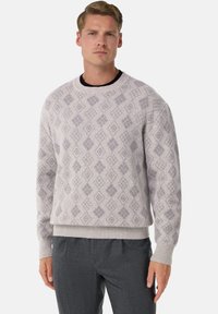 Man som bär en ljusgrå tröja med ett subtilt diamantmönster och mörkgrå byxor, stående mot en enfärgad vit bakgrund.