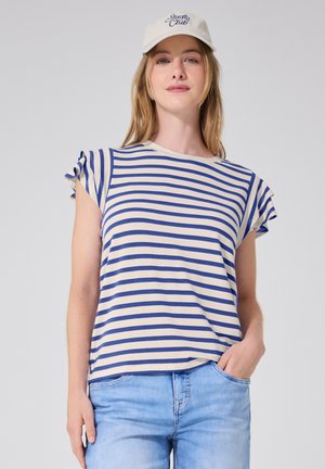 Joven mujer con una gorra blanca, camiseta de manga corta a rayas blancas y azules, y vaqueros azul claro, con una mano en el bolsillo, frente a un fondo liso.
