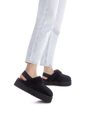 Zapatos de plataforma negros hechos de gamuza y piel de animal, que cuentan con una banda acolchada en la parte superior y una correa elástica en la parte posterior, llevados con jeans claros.