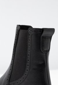 Musta nahkainen nilkkurit, joissa on sileä pinta, joustava sivupaneeli ja koristeelliset rei'itykset reunan varrella. Varustettu vedettävällä lenkillä.