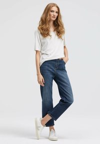 Weißes kurzärmliges Oberteil, lockere blaue Jeans mit geradem Schnitt und weiße Turnschuhe mit dezentem Branding vor einem hellen Hintergrund.