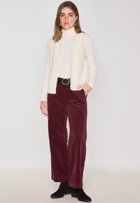 Gilet sans manches en polaire ivoire avec bordure en tricot, associé à un pantalon large en velours côtelé bordeaux taille haute. Ceinture noire avec boucle ronde.