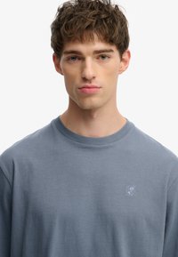 Camisa de algodón azul de manga larga con escote redondo y sutil logo bordado en el pecho. Textura suave y ajuste relajado.