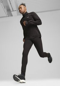 Juodas sportinis apranga, įskaitant ilgomis rankovėmis marškinėlį ir prigludusias leggingus. Marškinėliai turi aukštą apykaklę ir angas nykščiams, derinami su elegantiškais juodais sportbačiais.