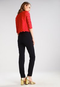 Top corto de encaje rojo con patrones florales, combinado con pantalones ajustados negros y sandalias de tacón doradas. Texturas suaves, colores contrastantes.