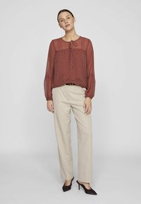 Rostfärgad transparent blus med prickar, slipskrage och långa puffärmar, ihop med beige skräddarsydda byxor och mörka klackar.