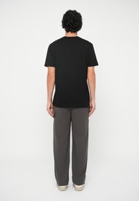 Persona con capelli ricci che indossa una maglietta nera a maniche corte, pantaloni larghi grigi con tasca laterale e sneakers bianche, in piedi di spalle.