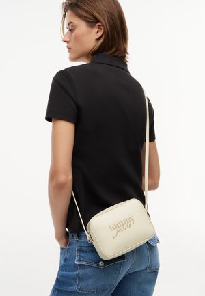 Mujer con una camiseta polo negra y vaqueros azules lleva un bolso bandolera crema con "tommy jeans" grabado en el frente.