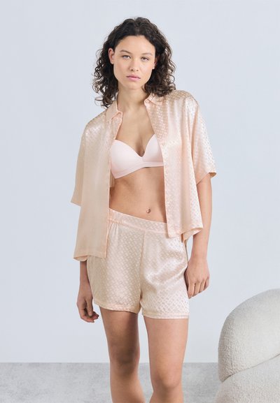 Femme portant une chemise en satin rose pâle à manches courtes avec des boutons, un short assorti et un soutien-gorge rose clair, debout dans une pièce minimaliste.