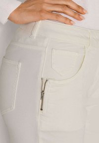 Weiße Denim-Jeans mit eng anliegendem Schnitt verfügen über eine Reißverschlusstasche und eine aufgenähte Pattentasche. Der Stoff hat eine glatte Textur.