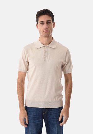 Giovane uomo con capelli scuri che indossa una polo beige a maniche corte e jeans blu, in piedi davanti a uno sfondo chiaro e uniforme.