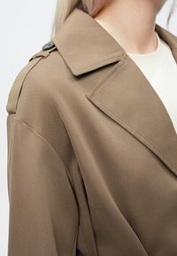 Taupefarbener Trenchcoat mit strukturiertem Kragen, glatter Textur und Knopfdetail auf der Schulter, teilweise sichtbar vor hellem Hintergrund.