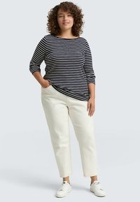 Dolga majica z dolgimi rokavi s črtami v mornarsko modri in krem barvi, kombinirana s kremasto barvnimi hlačami, dolgimi do gležnjev, ter belimi supergami. Model stoji z rokami v žepih.