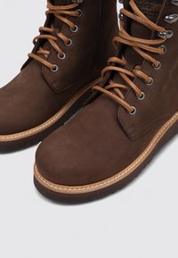 Botas de cuero marrón con una textura suave, que cuentan con cordones marrones y Ojetes de metal, una suela blanca cosida y un detalle lateral con cremallera.