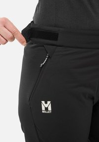 Les pantalons de randonnée Black Millet présentent une taille plate avec une sangle à fermeture auto-agrippante, une poche zippée et un logo argenté sur le côté.