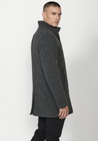 Abrigo gris de doble pecho con un alto cuello, elaborado en un tejido texturizado y con dos bolsillos laterales, exhibido en un maniquí de perfil.