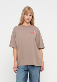 Smeđa oversize majica od pamuka s okruglim izrezom. Ima crveni Levi's logo na lijevoj strani prsiju. Opusten kroj i kratki rukavi.