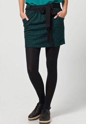 Jupe à motif vert avec une ceinture noire à nouer, deux poches, portée avec des collants noirs et des chaussures à lacets.