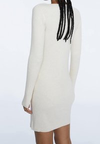 Robe tricotée blanche ajustée à rayures avec manches longues, présentant un col rond et des fentes sur les côtés pour faciliter le mouvement. Texture lisse.