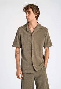 Camicia khaki a maniche corte in tessuto strutturato con colletto, chiusura frontale a bottoni e leggero lucido. Presenta cinture arrotolate e una vestibilità rilassata.