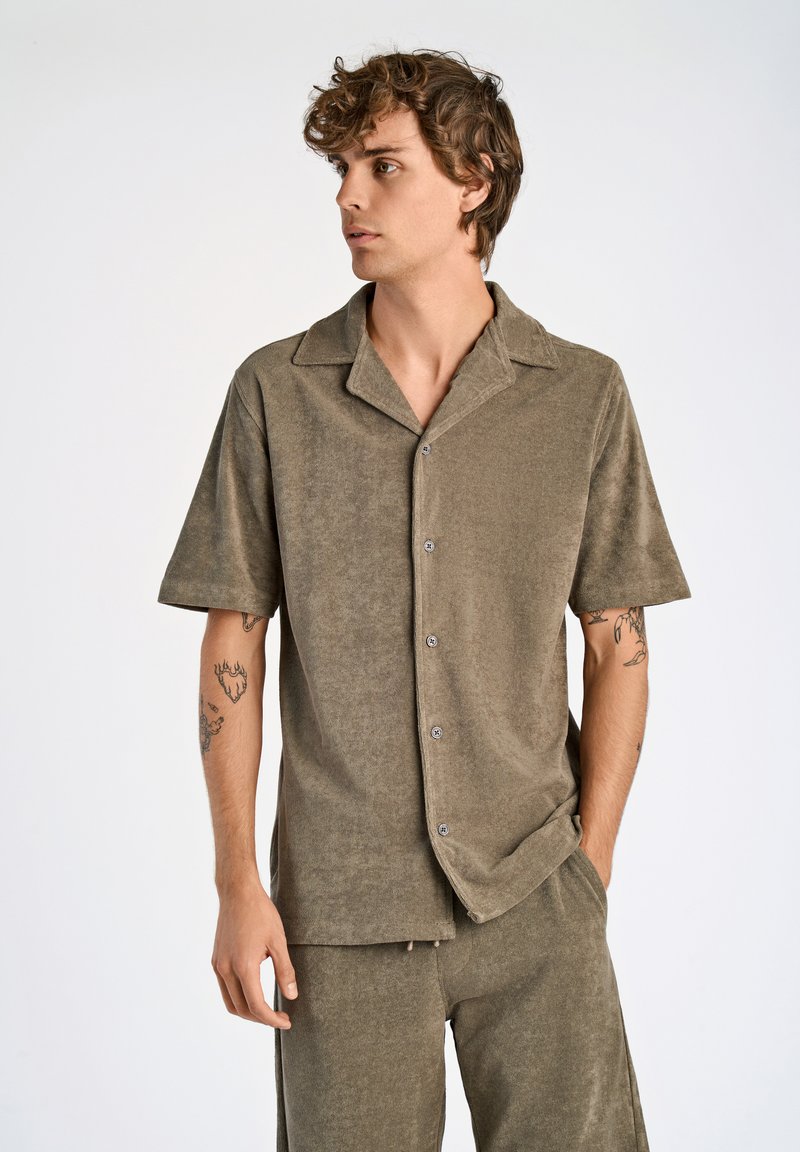 Camicia khaki a maniche corte in tessuto strutturato con colletto, chiusura frontale a bottoni e leggero lucido. Presenta cinture arrotolate e una vestibilità rilassata.