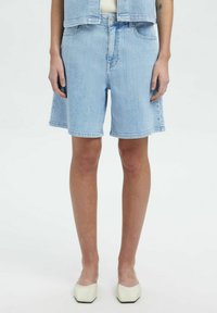 Pantalones cortos de mezclilla azul claro con un corte relajado, que presentan una cintura alta, dos bolsillos frontales y un dobladillo sin acabar, combinados con zapatos blancos.