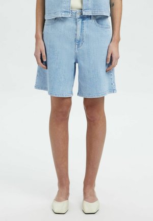 Shorts en denim bleu clair avec une coupe décontractée, taille haute, deux poches avant et un ourlet brut, associés à des chaussures blanches.