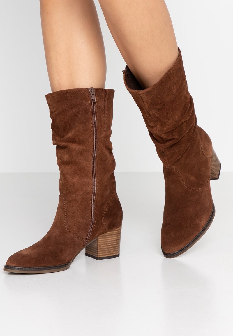 Botas de ante marrón hasta la pantorrilla con cremallera lateral y tacón bloque apilado, usadas en piernas contra un fondo blanco liso.