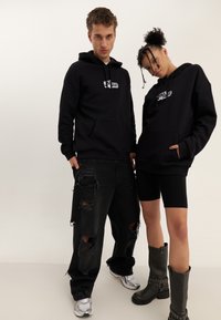 Čierne mikiny s prednými klokankami, na ktorých je vyobrazené logo. Čierne poškodené džínsy a čierne vysoké čižmy dokončujú outfit.