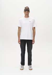 Camiseta blanca de algodón con un pequeño logo, combinada con jeans de mezclilla oscura y zapatillas ligeras. Un gorro de punto marrón completa el look.