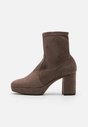 Brązowe zamszowe botki za kostkę z zaokrąglonym noskiem, wysokim słupkowym obcasem i elastycznymi bokami. Charakteryzują się gładką teksturą i minimalnymi detalami szycia.