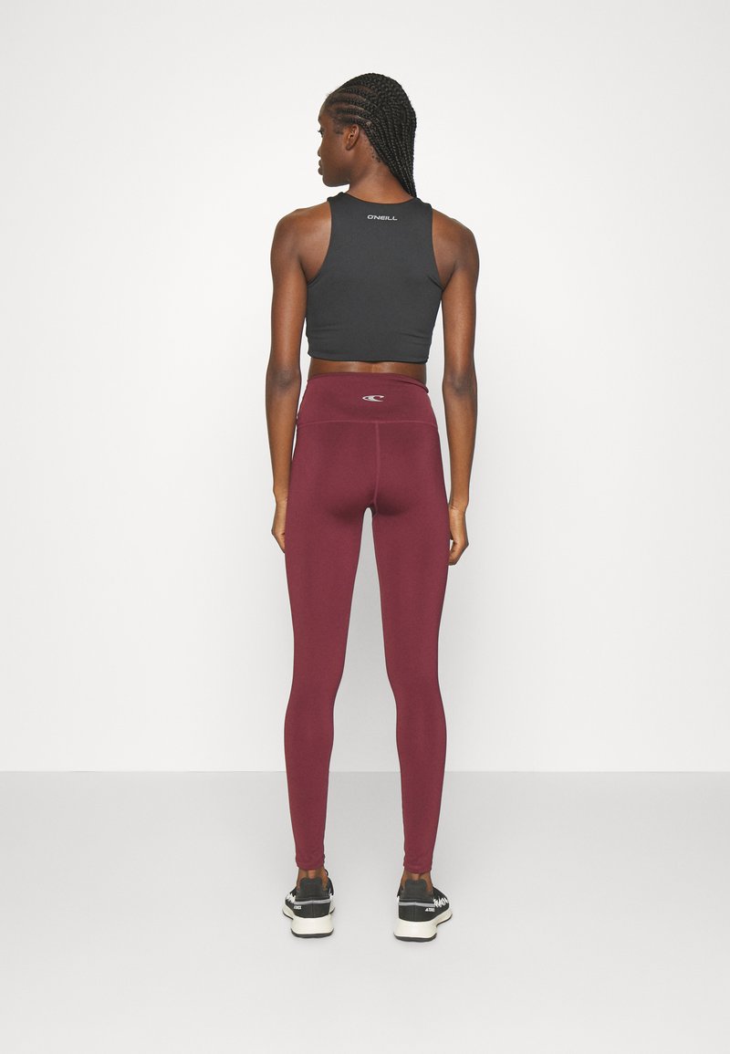 Schwarzes, kurzes Tanktop und bordeauxrote Leggings mit hoher Taille, aus elastischem Stoff; das Tanktop hat ein Logo auf der Rückseite, die Leggings haben einen subtilen Glanz.